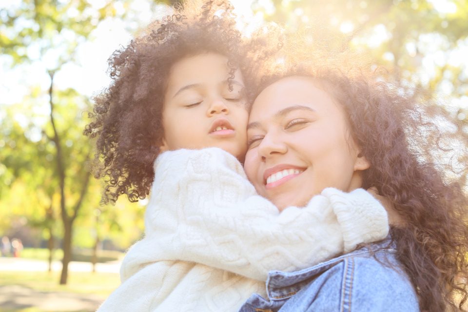 A mother laughing tenderly while embracing her young daughter who wears glasses, outdoors in warm sunlight.